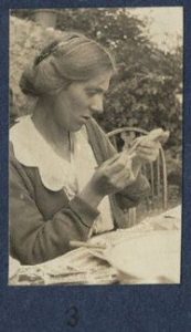 Fredegond Shove in a black and white photo taken by Lady Ottoline Morrell. She is sitting at a table in a garden and focussed on something in her hands.